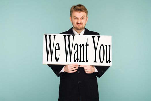 Angry beard young business man showing signboard with sign We Want You, on blue background