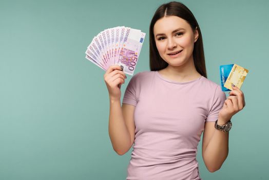Photo of caucasian brunette woman 20s smiling while holding credit card and fan of euro cash isolated over blue background - Image