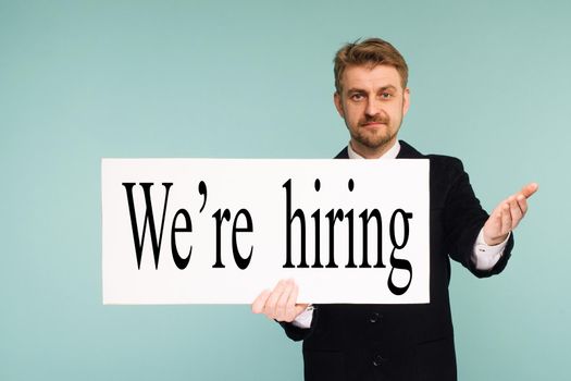 Happy smiling young business man showing signboard with sign we hiring, on blue background