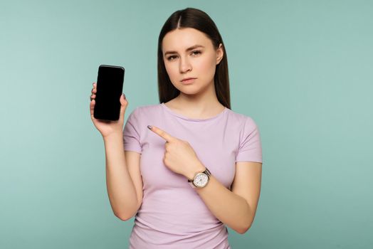 Young sad girl points at smartphone on blue background. The concept of Internet dependence on social networks
