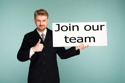 Happy smiling young business man showing signboard with sign Join Our Team thumbs up, on blue background