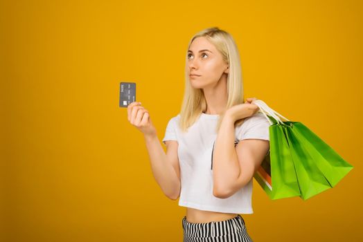 Close-up portrait of happy young beautiful blonde woman holding credit card and green shopping bags, isolated on yellow background - Image