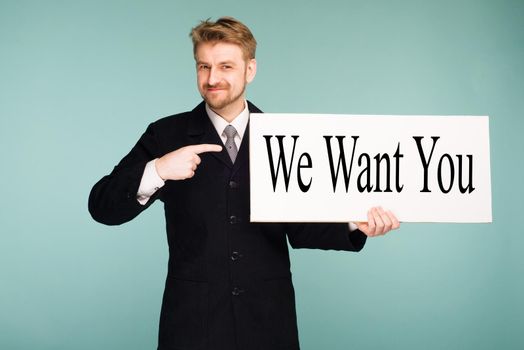 Happy smiling young business man points finger signboard with sign We Want You, on blue background