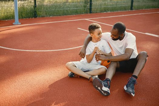 Young father and his son sitting on basketball court near the park. Man and boy wearing white t-shirts. They are talking and holding an orange ball.