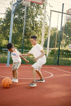 Two multiracional brothers playing basketball on a court near the park. Boys wearing white t-shirts. Older brother teach little one how to play basketball.