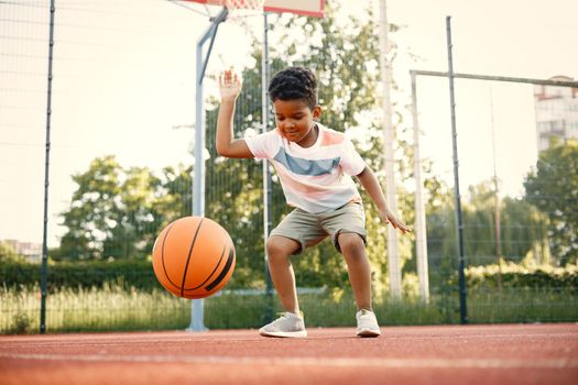 Young boy standing on basketball court near the park. Boy wearing white t-shirt. Boy playing with basketball ball.