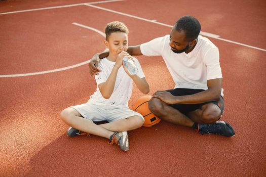 Young father and his son sitting on basketball court near the park. Man and boy wearing white t-shirts. They hugging and boy drinking a water.