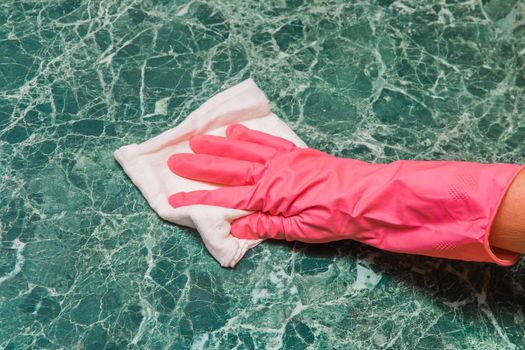 Housewife's hand in a pink household glove wipes a green marble surface with a white cloth in the kitchen.