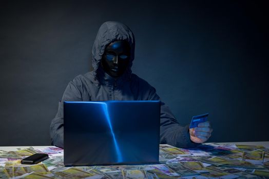 Anonymous hacker in mask programmer uses a laptop to hack the system in the dark. The concept of cybercrime theft of money from bank cards