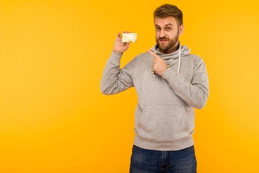 Attractive man in a gray hoodie points a finger at the credit card that is holding in his hand on a yellow background - image