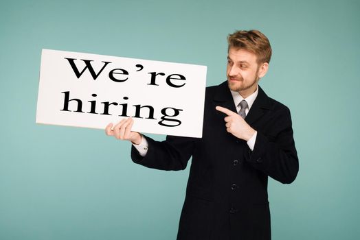 Happy smiling young business man points finger on signboard with sign we hiring, on blue background
