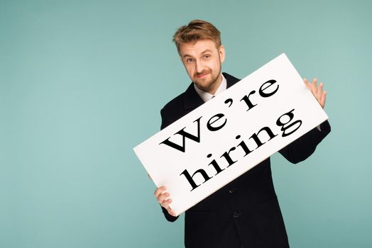 Happy smiling young business man showing signboard with sign we hiring, on blue background