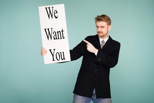 Happy smiling young business man points finger signboard with sign We Want You, on blue background