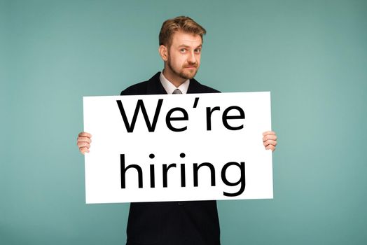 Happy smiling young business man showing signboard with sign we hiring, on blue background