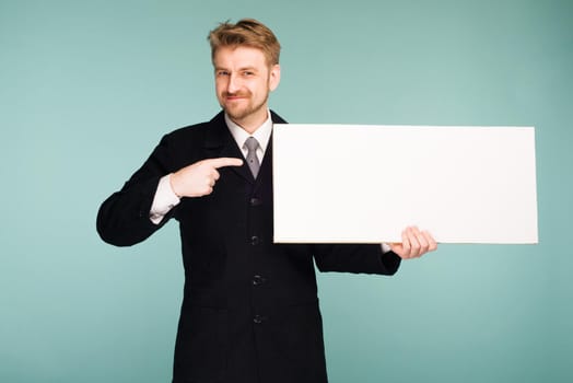 Happy smiling young business man points finger blank signboard, on blue background