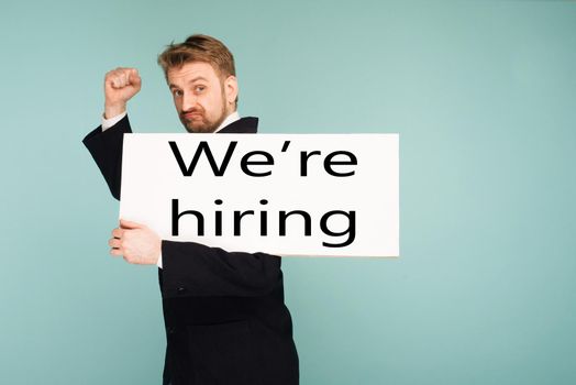 Funny young business man showing signboard with sign we hiring, on blue background