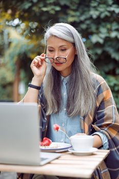 Concentrated mature Asian lady with glasses and strawberry looks at laptop screen at table on outdoors cafe terrace on autumn day