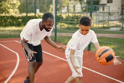 Young father and his son playing basketball in basketball court near the park. Man and boy wearing white t-shirts. Boys holding an orange basketball ball with black stripes.l