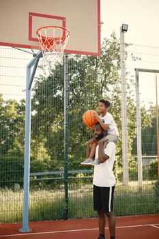 Black father holding on shoulders his little son to help him to score a basket. They standing on a basketball court. Man and boy wearing white t-shirts.