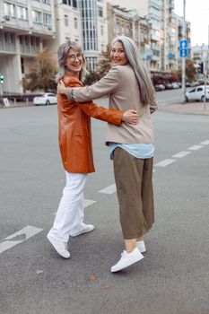 Smiling mature Asian lady and grey haired companior hug walking along modern city street on autumn day backside view