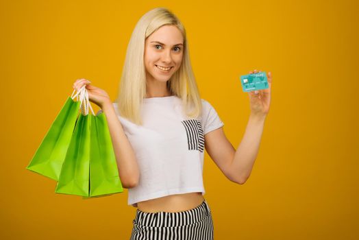 Close-up portrait of happy young beautiful blonde woman holding credit card and green shopping bags, looking at camera, isolated on yellow background - Image