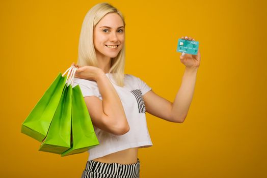 Close-up portrait of happy young beautiful blonde woman holding credit card and green shopping bags, looking at camera, isolated on yellow background - Image