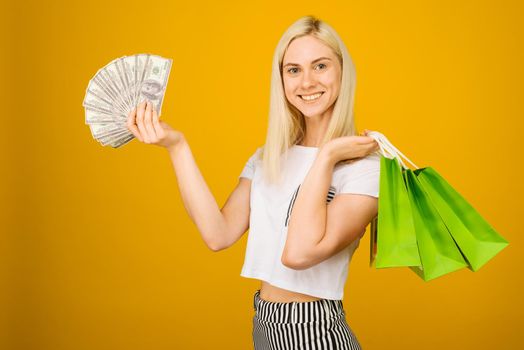 Close-up portrait of happy young beautiful blonde woman holding money and green shopping bags, looking at camera, isolated on yellow background - Image