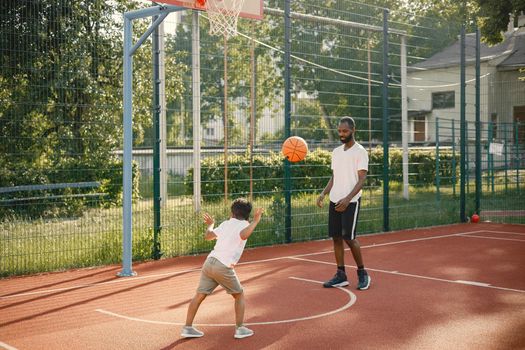 Young father and his son playing basketball in basketball court near the park. Man and boy wearing white t-shirts. Boys holding an orange basketball ball with black stripes.l