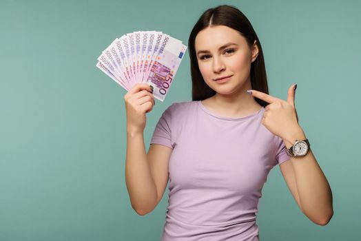 Portrait of a happy girl pointing finger at bunch of money banknotes isolated over blue background - Image.