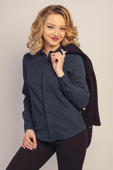 Portrait of smiling beautiful young business woman standing with hand holding jacket behind her shoulder against grey background and posing fashionable. Looking at camera.