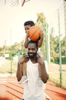 Black father holding on shoulders his little son to help him to score a basket. They standing on a basketball court. Man and boy wearing white t-shirts.