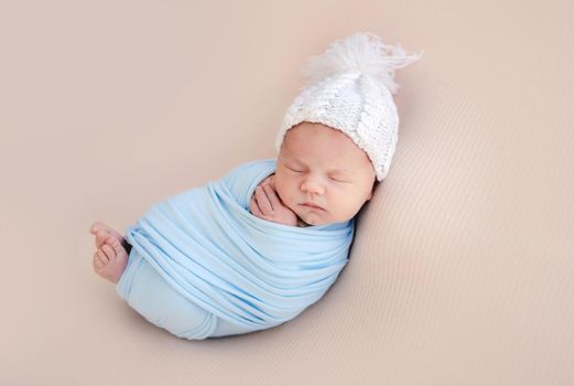 Newborn baby boy swaddled in blue fabric and wearing hat sleeping. Infant child kid napping during studio photoshoot