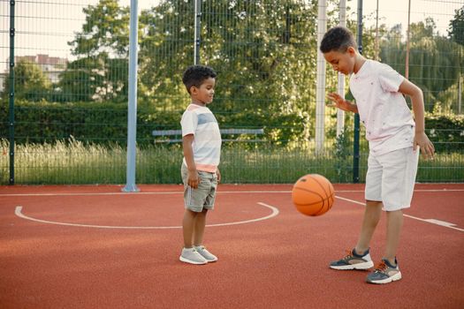 Two multiracional brothers playing basketball on a court near the park. Boys wearing white t-shirts. Older brother teach little one how to play basketball.