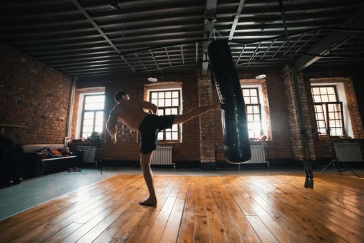 An athletic man boxer training with a punching bag in the gym. Kicking the bag with a leg. Mid shot