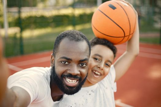 Young father and his son standing on basketball court near the park. Man and boy wearing white t-shirts. Father and son make a selfie.