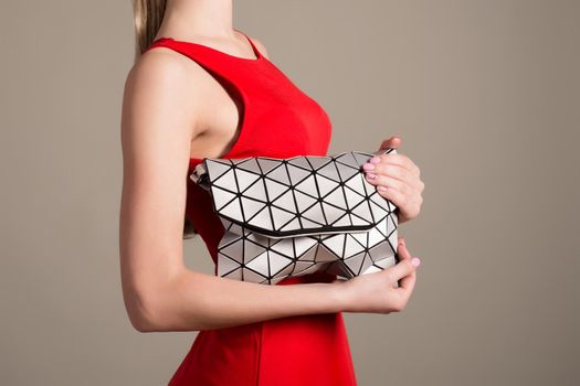 Attractive slim girl in a red evening dress holds a stylish bag of silvery triangles on a gray background