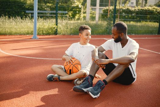 Young father and his son sitting on basketball court near the park. Man and boy wearing white t-shirts. They are talking and holding an orange ball.