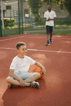 Young boy sitting on basketball court near the park. Boy wearing white t-shirt. There are blurred african man behind.