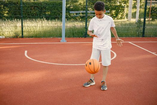 Young boy standing on basketball court near the park. Boy wearing white t-shirt. Boy playing with basketball ball.