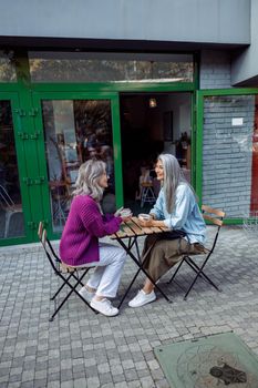 Positive mature woman and grey haired Asian companion with tasty coffee spend time together at table on outdoors cafe terrace on autumn day