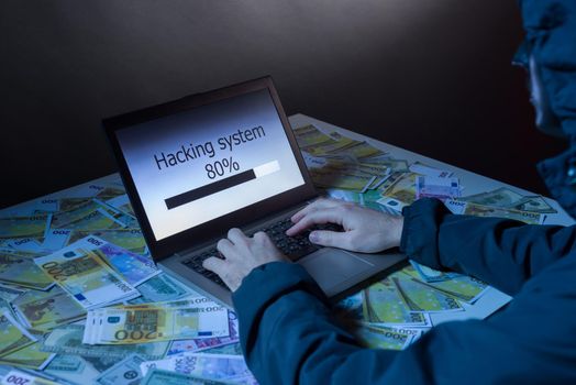 Anonymous hacker programmer uses a laptop to hack the system in the dark. The concept of cybercrime and hacking database. Money is scattered on the table. Focus on laptop