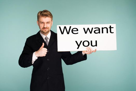 Happy smiling young business man showing signboard thumbs up with sign We Want You, on blue background