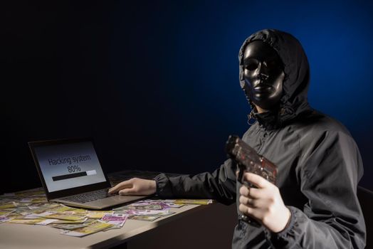 Anonymous hacker programmer uses a laptop holding a gun in his hand to hack the system in the dark. Creation and infection of malicious virus. The concept of cybercrime. Focus on laptop