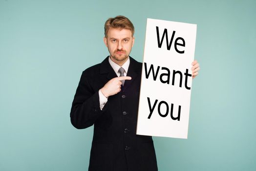 Happy smiling young business man points finger signboard with sign We Want You, on blue background