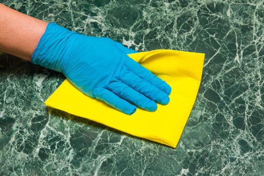 Housewife's hand in a blue latex glove wipes a green marble surface with a yellow napkin in the kitchen.