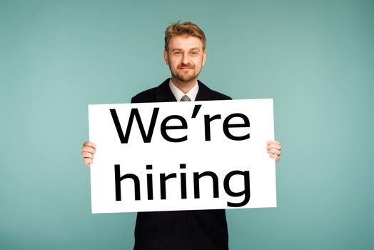 Happy smiling young business man showing signboard with sing we're hiring, on blue background