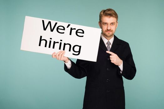 Happy smiling young business man points finger signboard with sing we're hiring, on blue background