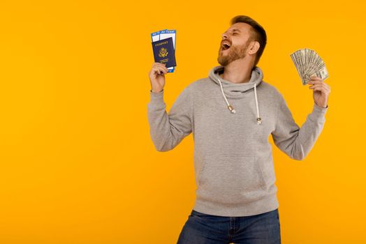 A handsome man in a gray hoodie rejoices in winning the lottery. He is holding a passport with air tickets and money dollars on a yellow background. - image