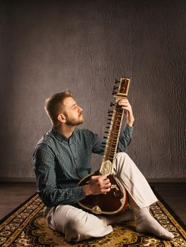 Portrait of a European man playing the sitar sitting on the carpet- Image