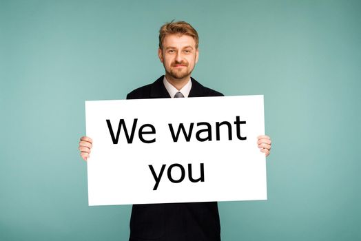 Happy smiling young business man showing signboard with sign We Want You, on blue background